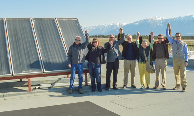 A line of men and women raise clasped hands in front of the Cranbrook mountains and a thermal solar panel.