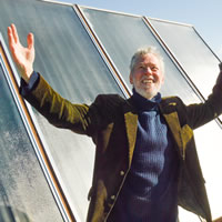 Guy Dauncey has white hair and a beard. He stands in front of solar panels with his arms spread wide.