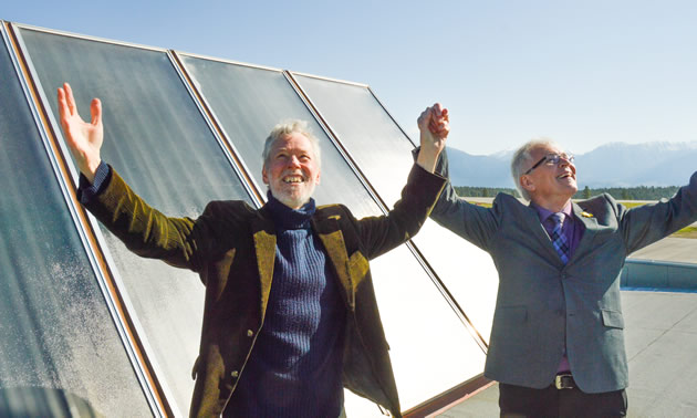 Guy Dauncey and Mayor Wayne Stetski pose in front of solar panels with their arms spread wide.