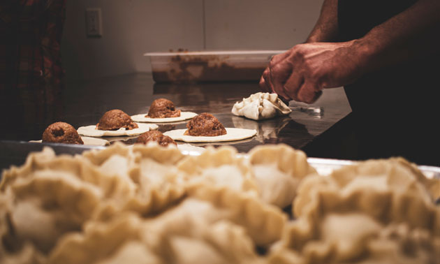 Close-up of person hand-crafting empanadas, with tray of made empanadas in foreground. 