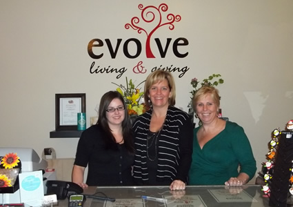 Leanne stands between her two employees behind the counter in front of the Evolve Living and Giving logo.