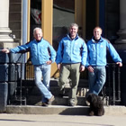 Two owners and the general manager of Everything Revelstoke stand on the steps of their new building. All wear sky-blue RMR jackets. A little black dog sits at their feet.