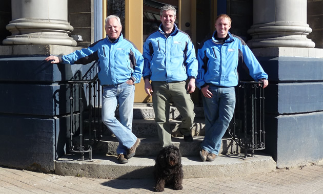 Two owners and the general manager of Everything Revelstoke stand on the steps of their new building. All wear sky-blue RMR jackets. A little black dog sits at their feet.