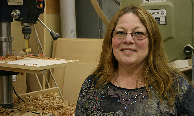 A woman in glasses stands beside woodworking equipment.