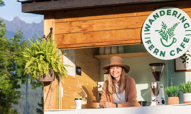 A woman is standing inside her pop-up cafe, with a fern hanging at the front.