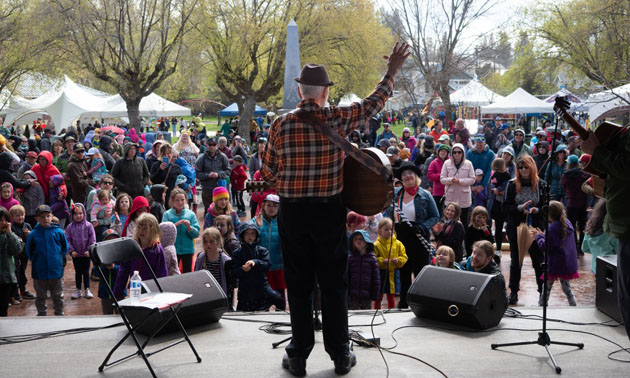 Fred Penner, standing on stage with back turned to camera, crowd of kids and people watching him. 