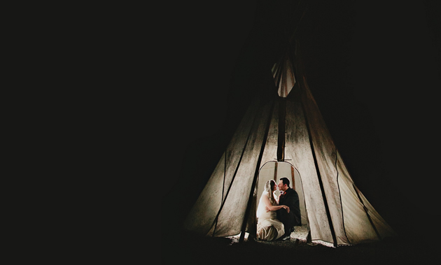 A bride and groom sit inside a lit tepee against an all-black background.