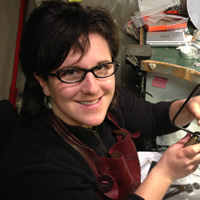 Arleigh has short brown hair and glasses, she sits at a curved and cluttered work dest, holding some of the tools of the trade.