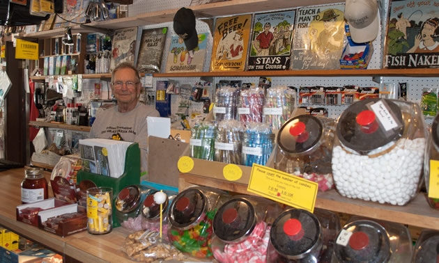 Lymbery stands behind a counter displaying penny-candy jars and a number of other colourful wares.