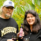 Casually dressed man and woman hold a small bottle and two wine glasses holding a dark red liquid.