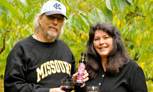 Casually dressed man and woman hold a small bottle and two wine glasses holding a dark red liquid.