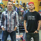Two casually dressed young men stand, hands in pockets, before an indoor array of ski and snowboard equipment.