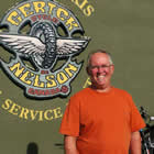 Broadly smiling man in an orange T-shirt in foreground, with elaborate business logo for Gerick Cycle and Ski on the wall behind him.