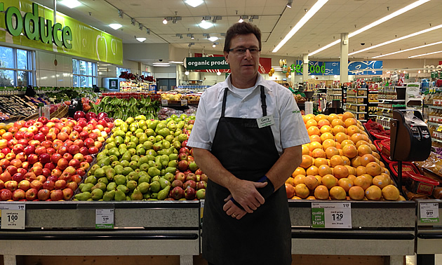 Man wearing white shirt and brown bib apron stands in front of fully-stocked bins of fruit in a store's produce department.