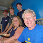 Smiling man in foreground, seated beside young woman, with three young men in background, in stadium broadcast booth.