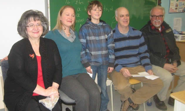 Five people in a row, including two women and two men, seated, and one boy, standing