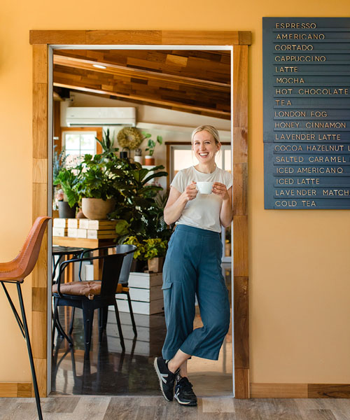 Women leaning against doorway holding cup of coffee and smiling. 