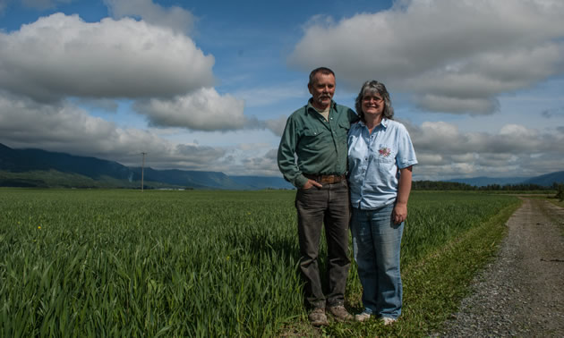 Roy and Sherry stand with their arms around each other beside a dirt road. Beside them is a green field and a blue sky with puffy clouds.