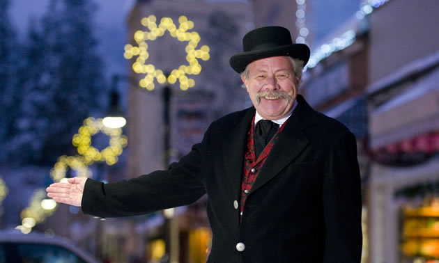 Broadly smiling man wearing  a black bowler hat and topcoat gestures with his right hand toward the festive lights on the street behind him
