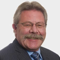 A head shot of mayor Greg Granstrom. He has grey hair, a moustache and glasses, wearing a grey suit with blue shirt and tie. 