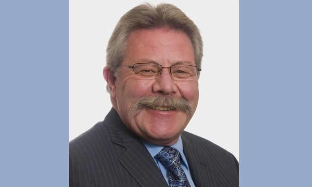 A head shot of mayor Greg Granstrom. He has grey hair, a moustache and glasses, wearing a grey suit with blue shirt and tie. 