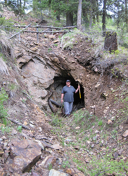 A man in jeans holding a pickaxe stands in front of a huge hole in a hillside. 