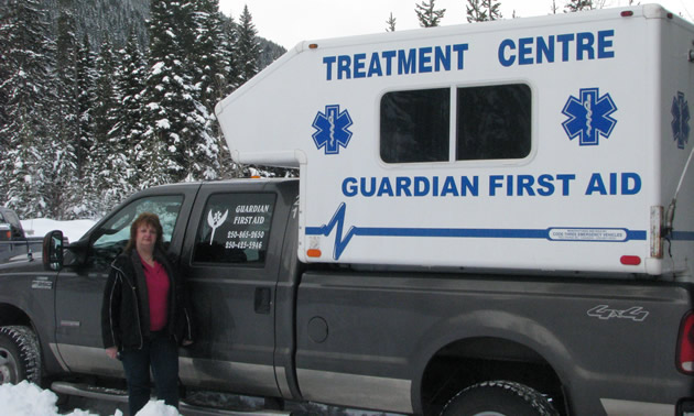 A woman standing beside a black pickup truck carrying a white camper with a sign saying Treatment Centre Guardian First Aid