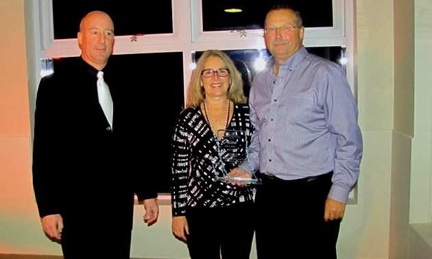 A couple hold a clear glass award while a presenter stands nearby.