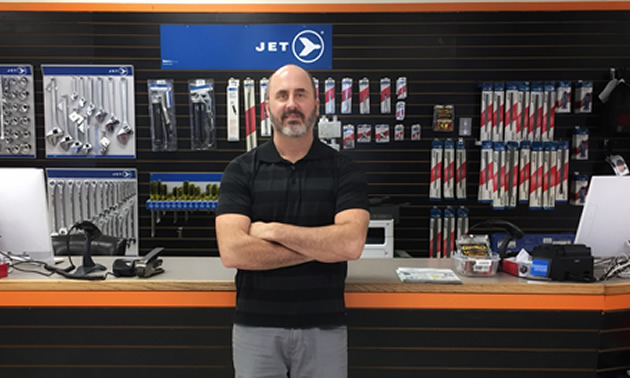 Mike Konkin, one of the owners of Trail Hammer and Bolt, is standing in front of the store counter with goods for sale in the back.