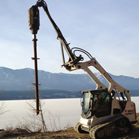 A small Bobcat machine holds a helical pile suspended above the ground, with lake and mountains in the background.