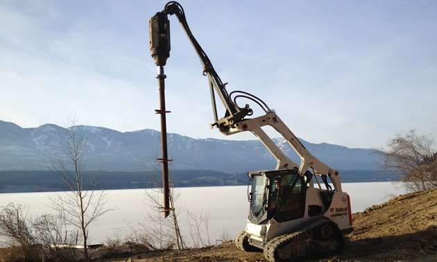 A small Bobcat machine holds a helical pile suspended above the ground, with lake and mountains in the background.