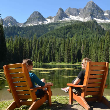 Two people sitting in wooden lawn chairs overlooking a small lake, forest and mountains