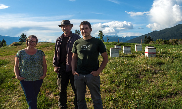 Jen Comer, Jim Comer, and Joel Comer stand against a brilliant sky and green field with bee boxes behind them.