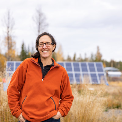 Dr. Ilona Hale standing in field on overcast day, row of solar panels in picture behind her. 