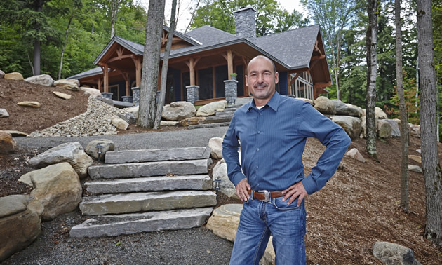 Man wearing blue shirt and jeans stands before stone steps leading up to a beautiful log home