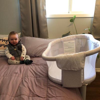 Sleep bassinet set up in hotel room, baby sitting on bed. 