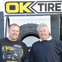 Two men, one younger and one older, stand under an OK Tire sign