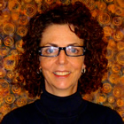 Joanne smiles at the camera in front of a gold-swirly pattern. She wears glasses and a dark blue shirt and has curly dark brown hair.