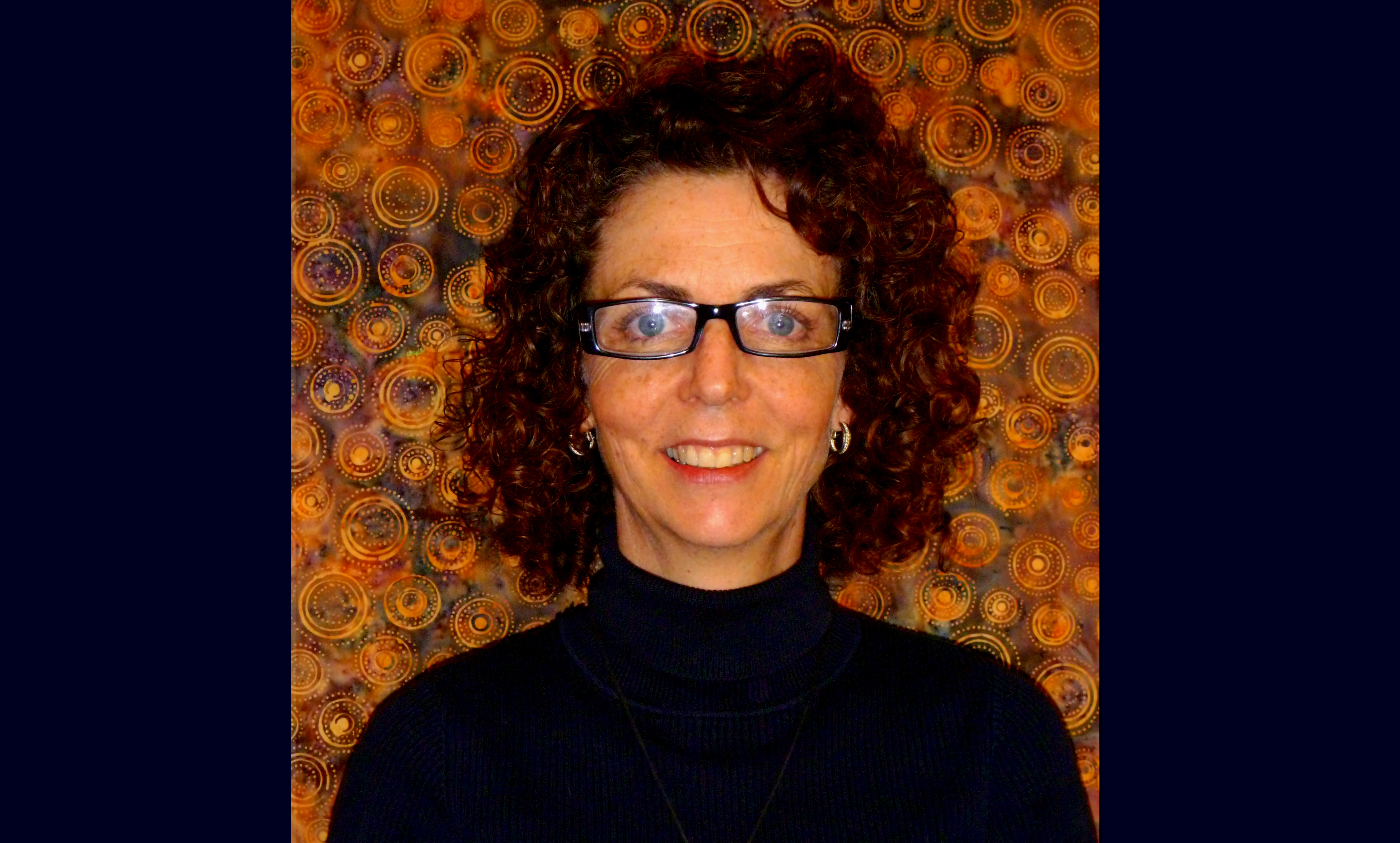 Joanne smiles at the camera in front of a gold-swirly pattern. She wears glasses and a dark blue shirt and has curly dark brown hair.