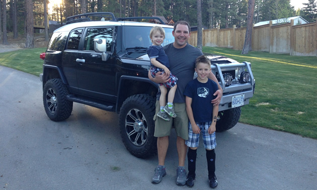 Smiling man with two young children stands in front of a rugged-looking vehicle