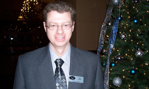 Man in business suit, wearing a Prestige Resorts name tag, stands beside a Christmas tree.