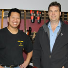 Two smiling men standing before a wall display of guitars