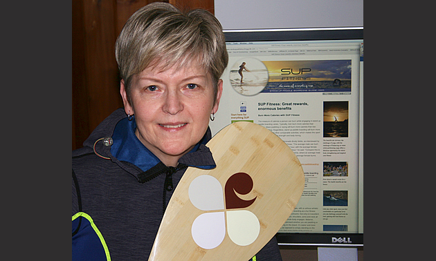 Kathy Verigin has short blonde hair and is holding a paddle. Behind her is a screen showing her new webpage.