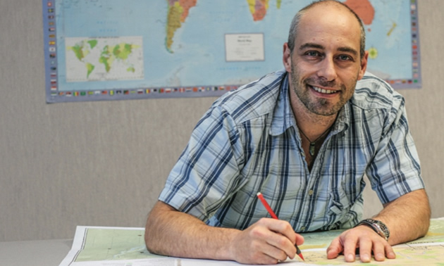 Picture of man wearing a striped shirt, sitting at a table and holding a pencil. 