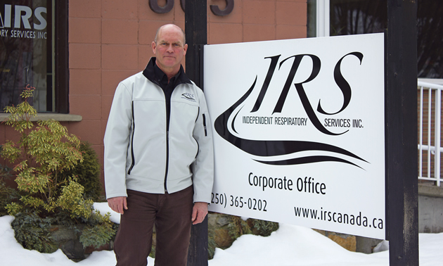 Balding man in a white winter jacket stands beside a large sign saying 