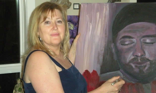 A blonde woman holds a paintbrush next to a work-in-art of a painting of a man.