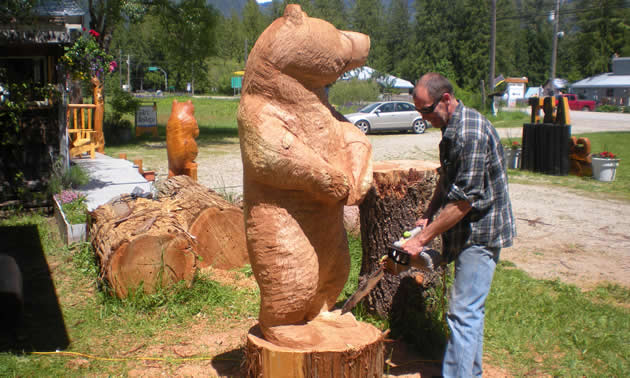 A man in a plaid shirt works on a life-sized carving of a bear with a chainsaw.
