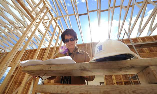 Camera points upward at a woman standing within a building being constructed, looking at plans; sun shines through the open rafters.