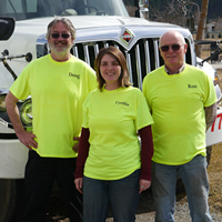 Cynthia Howard, manager of Kettle Valley Waste, stands with owner Ron Liddle and a driver. They all wear bright yellow company shirts.