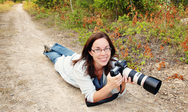 Smiling young woman holding a professional-style camera, lying prone on a pathway